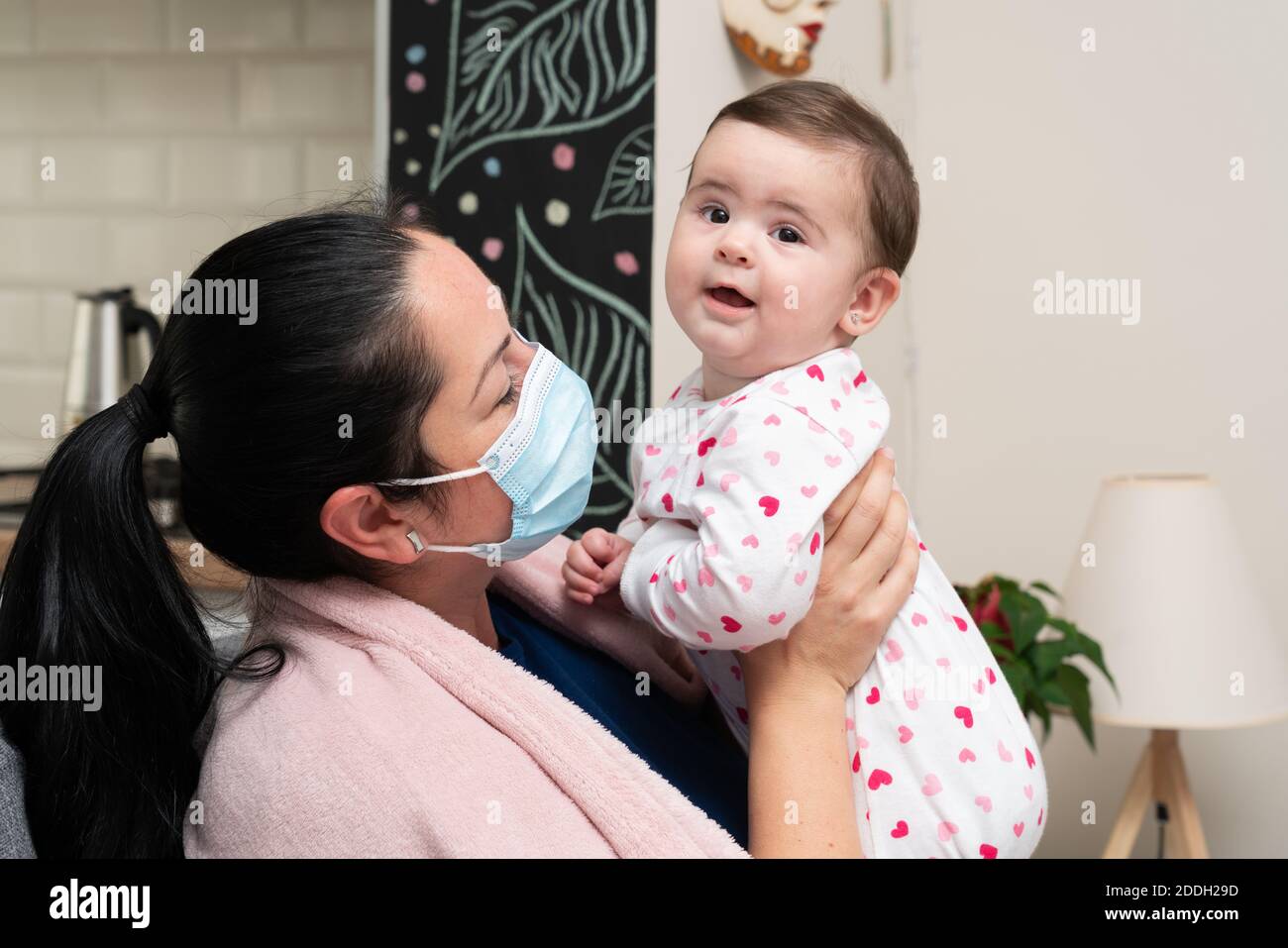 Adult woman model having sars flu covid19 illness symptoms covering nose and mouth using medical or surgical mask playing with baby girl daughter as p Stock Photo
