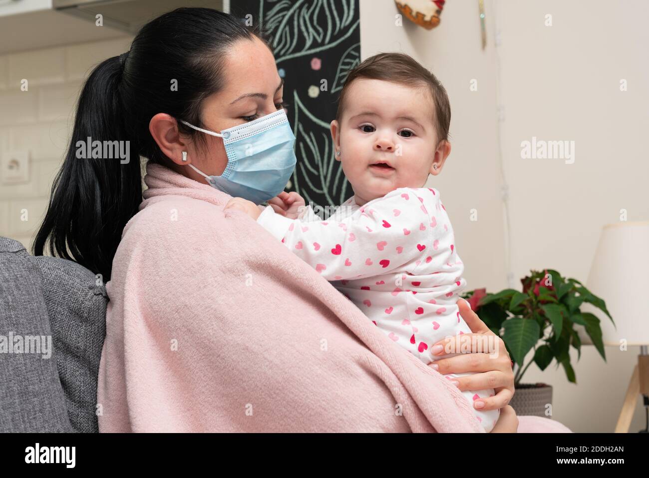 Ill female adult model wearing surgical or medical covid19 cold flu protective mask hugging cute baby child as pandemic family time concept Stock Photo