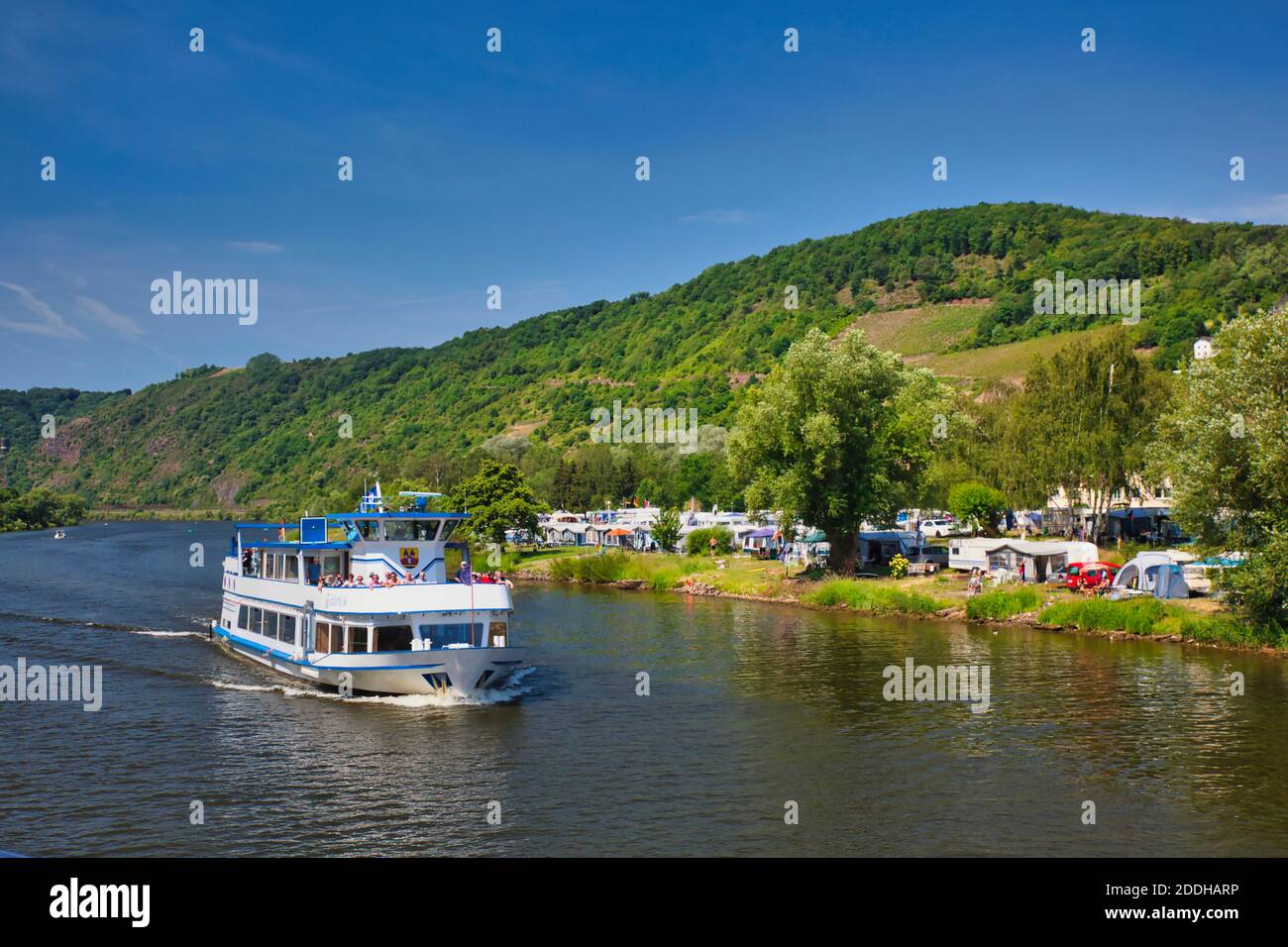 A cruise boat on the River Rhine in Germany passes by a large campsite on the river banks with hillside beyond Stock Photo
