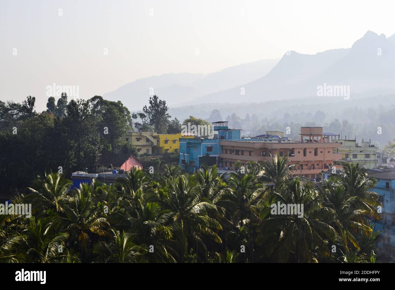 Palm trees and colourful houses against scenic morning view of mountains range in haze. Tropical landscape, Sulthan Bathery, Kerala, India. Stock Photo