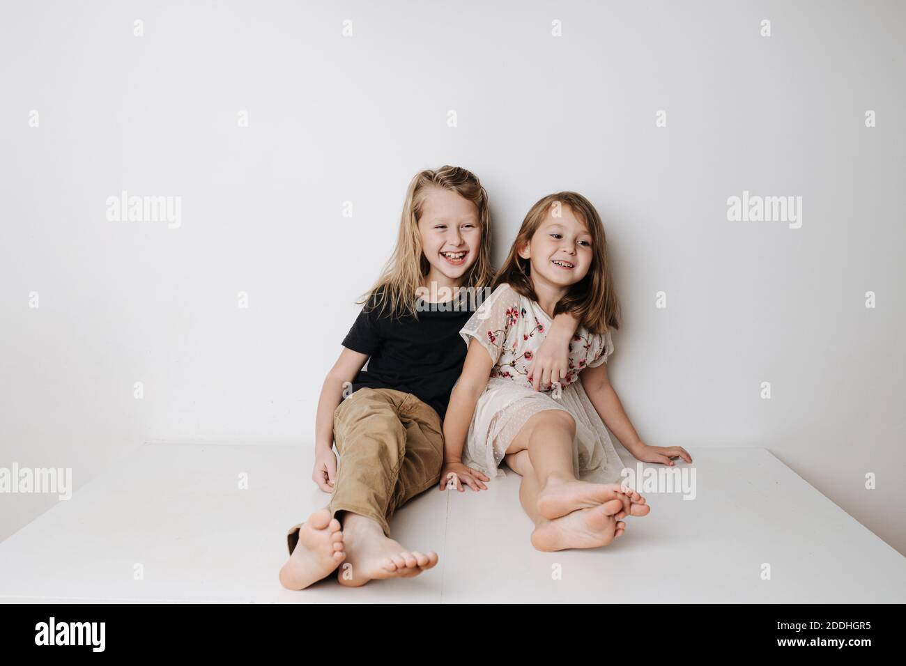 Smiling siblings sitting together on table. Boy hugs girl, she leans away a bit. Stock Photo