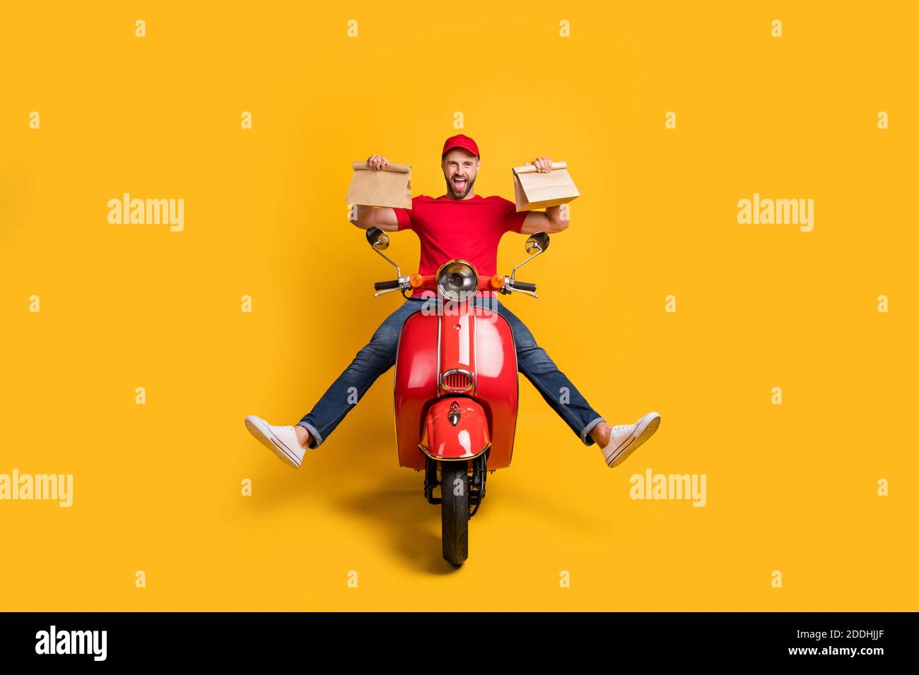 Photo of careless crazy guy drive bike hold two packages wear red tshirt cap jeans isolated