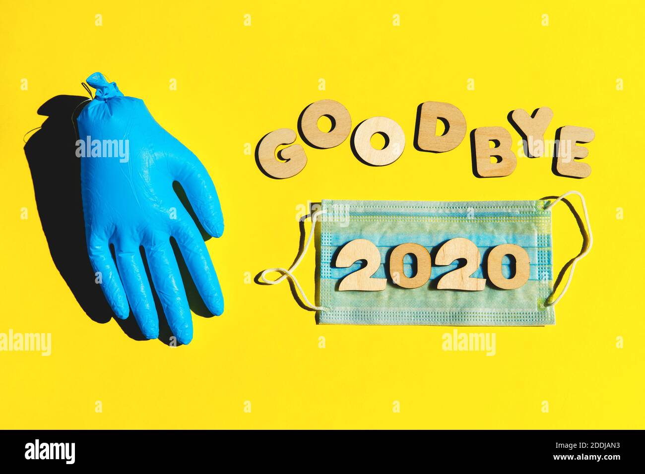 Words GOODBYE 2020 from wooden letters, inflated medical glove waving bye-bye and facemask on a yellow background. Year 2020 and epidemic concept. Stock Photo