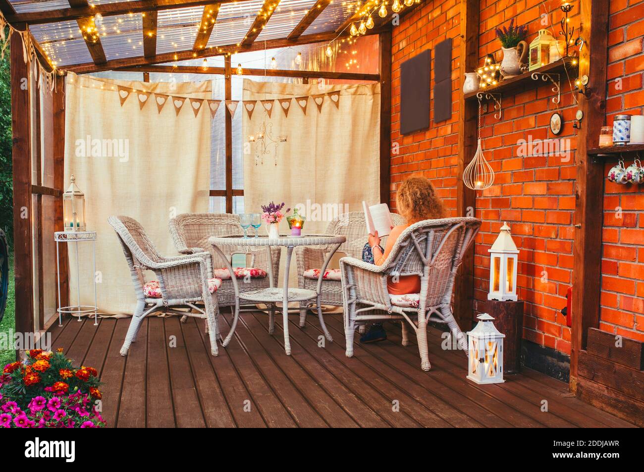 Hygge balcony concept. Woman reading a paper book on home balcony outdoors. Party string lights illuminating the space. Lot of candles and lanterns. Stock Photo
