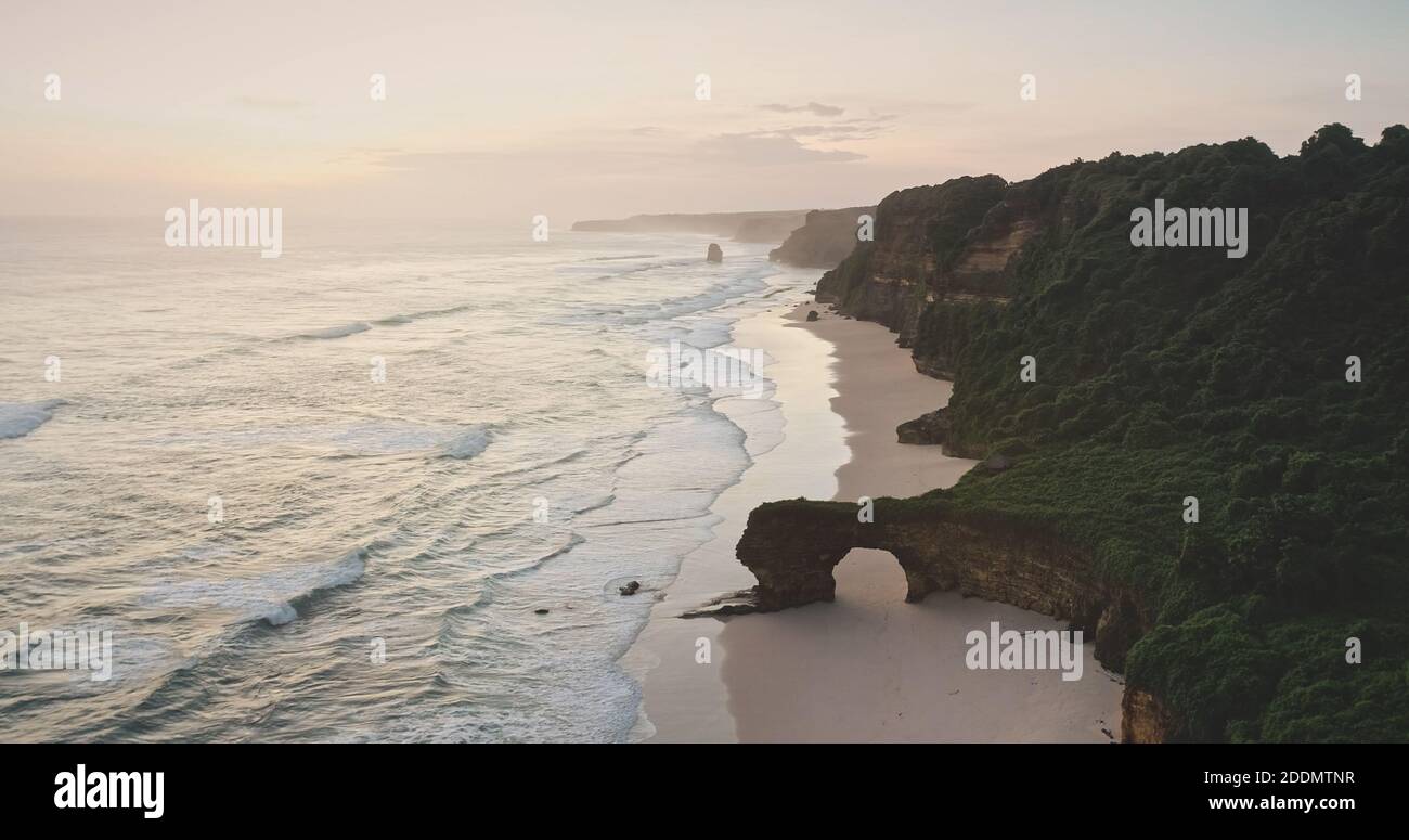 Sunrise at ocean cliff shore with giant hole at rock wall aerial view. Tropical nature landscape of paradise sand beach and green grass rocky coast. Tourist attraction of Sumba Island Stock Photo