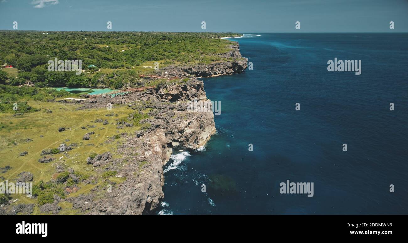 Cliff ocean coastline with green plants and trees at azure limpid lake. Indonesia attraction with building and bridge on rock sea shore of Sumba Island. Cinematic tropic landscape at drone shot Stock Photo