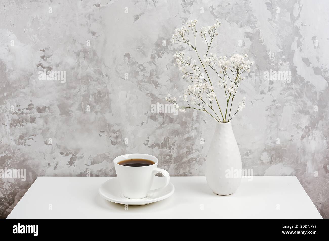 Cup of coffee and bouquet of small delicate flowers gypsophila in vase on white table agains gray stone wall. Copy space Minimal style. Concept Good m Stock Photo