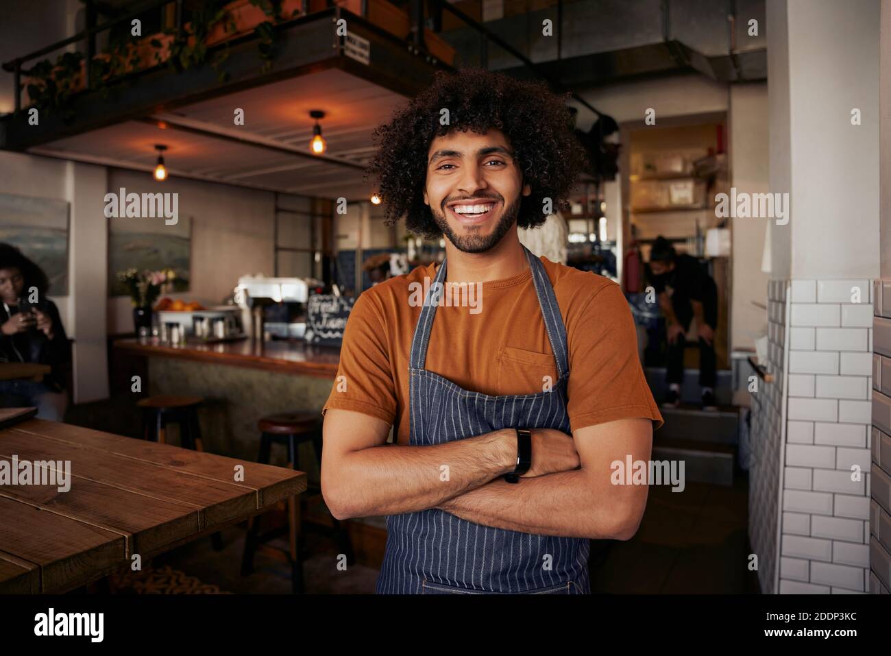 Handsome young male waiter wearing apron standing with crossed arms in cafe looking at camera Stock Photo