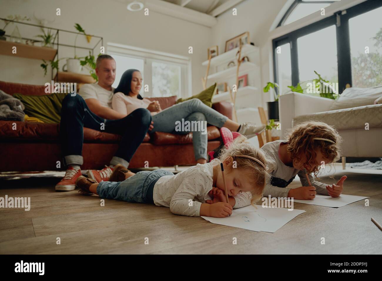 Parents sitting on couch with two cute beautiful daughters lying on floor creating artwork on paper at home Stock Photo