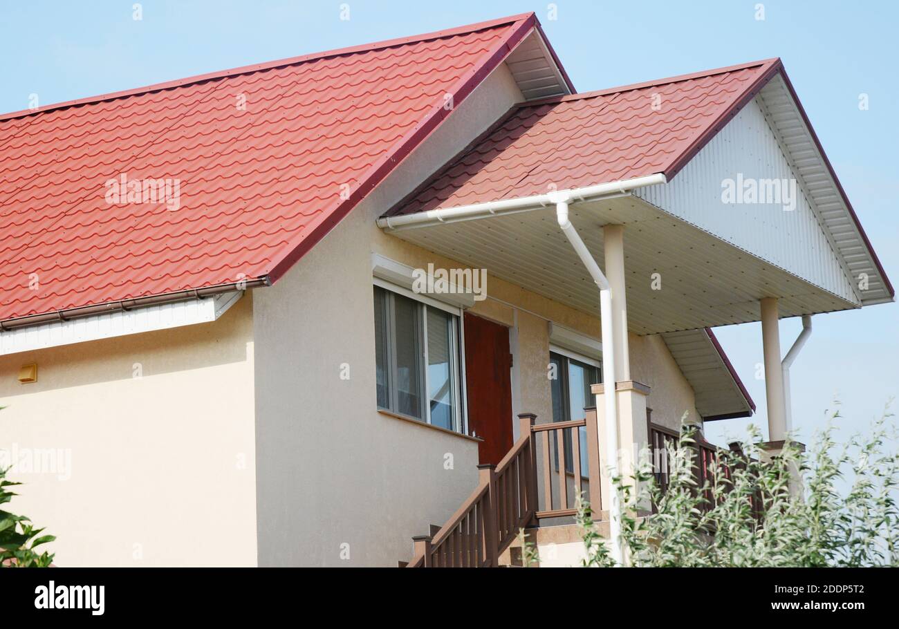 A close-up on a stucco house with a red metal tiled gabled roof with soffit and fascia boards, rain gutters with two downspouts and a covered porch wi Stock Photo