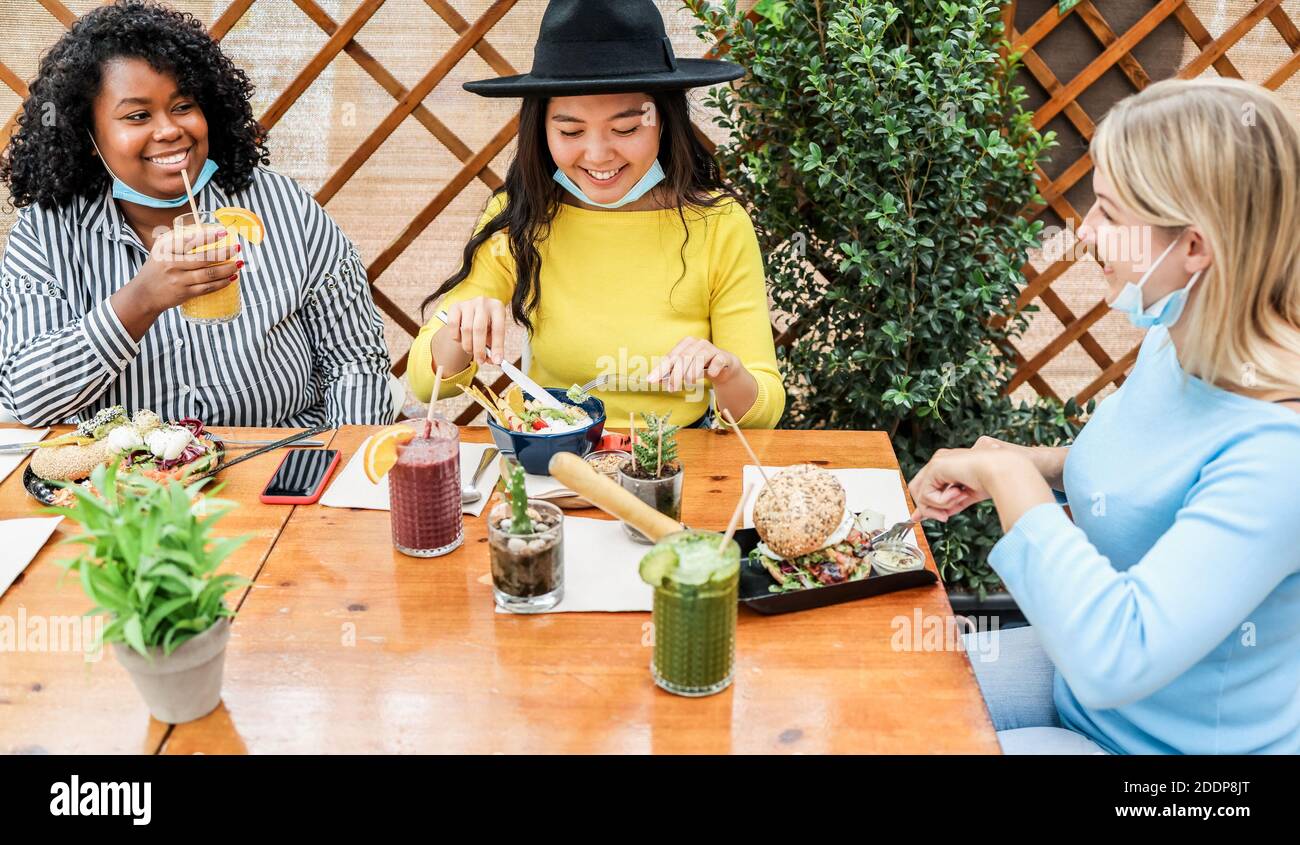 Multiracial young women eating brunch during coronavirus outbreak - Girls wearing protective masks at trendy restaurant - Healthy lifestyle and social Stock Photo