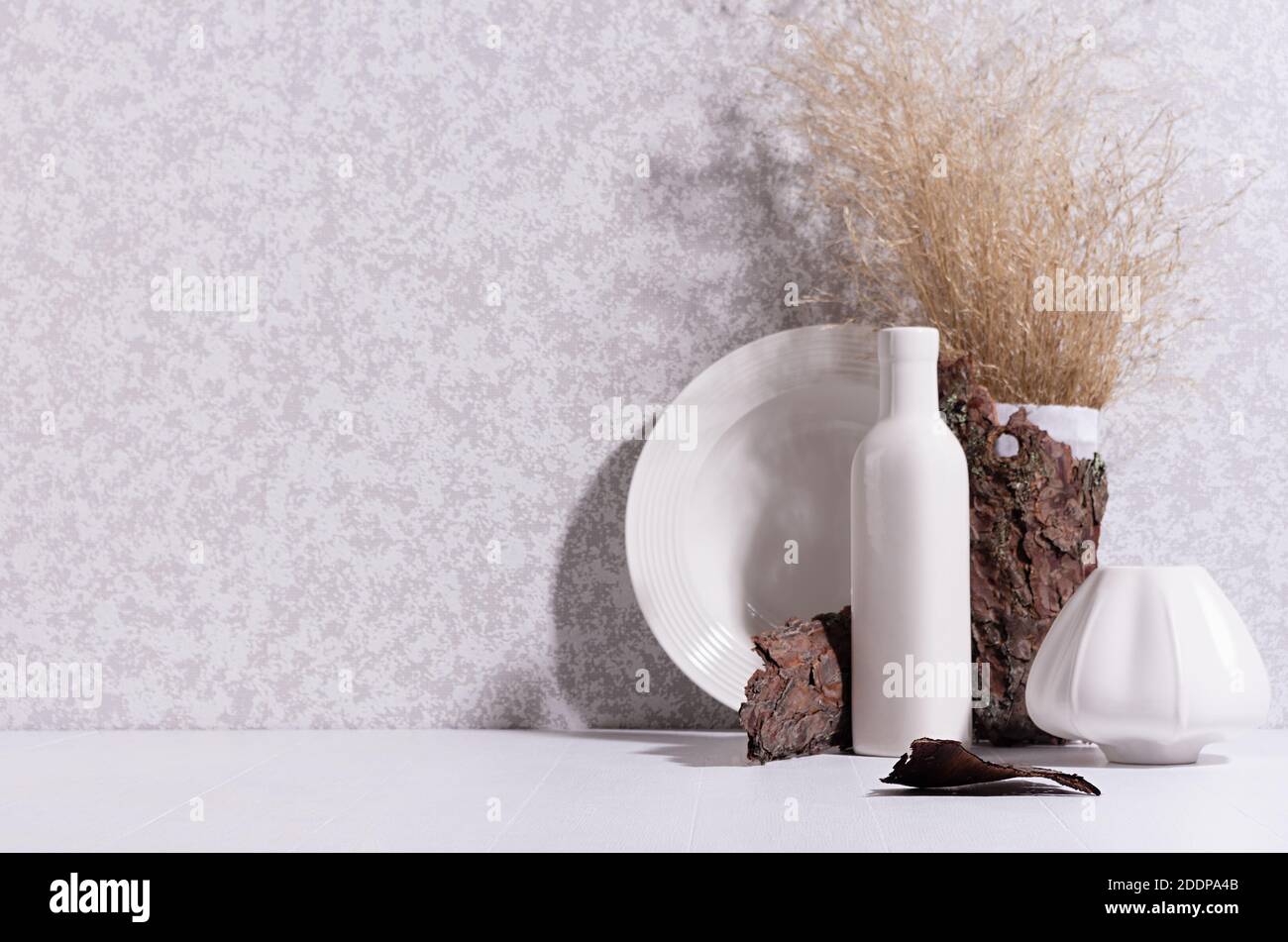 Scandinavian minimalist interior with ceramic vase, dish, dried grass bouquet, brown tree bark, moss in sun beam with shadow on white wood table. Stock Photo