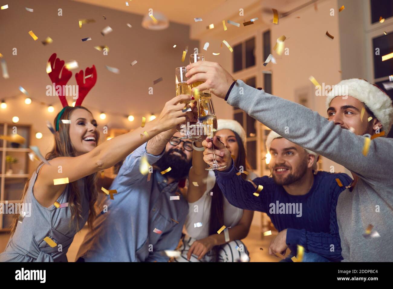 Group of young positive friends in decorative hats celebrating Christmas with champagne during party Stock Photo