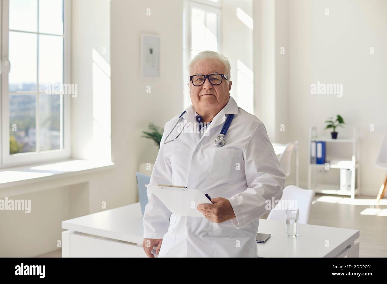 Experienced senior doctor standing in hospital office with clipboard in hand looking at camera Stock Photo