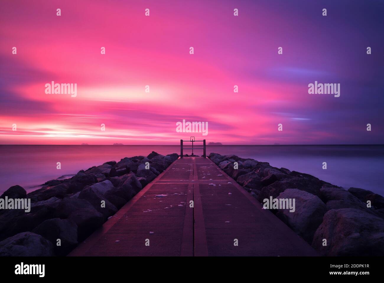 A long exposure captures a colourful sunrise as clouds streak through a pink and purple sky. The stone path on top of a groyne leads out into the sea. Stock Photo