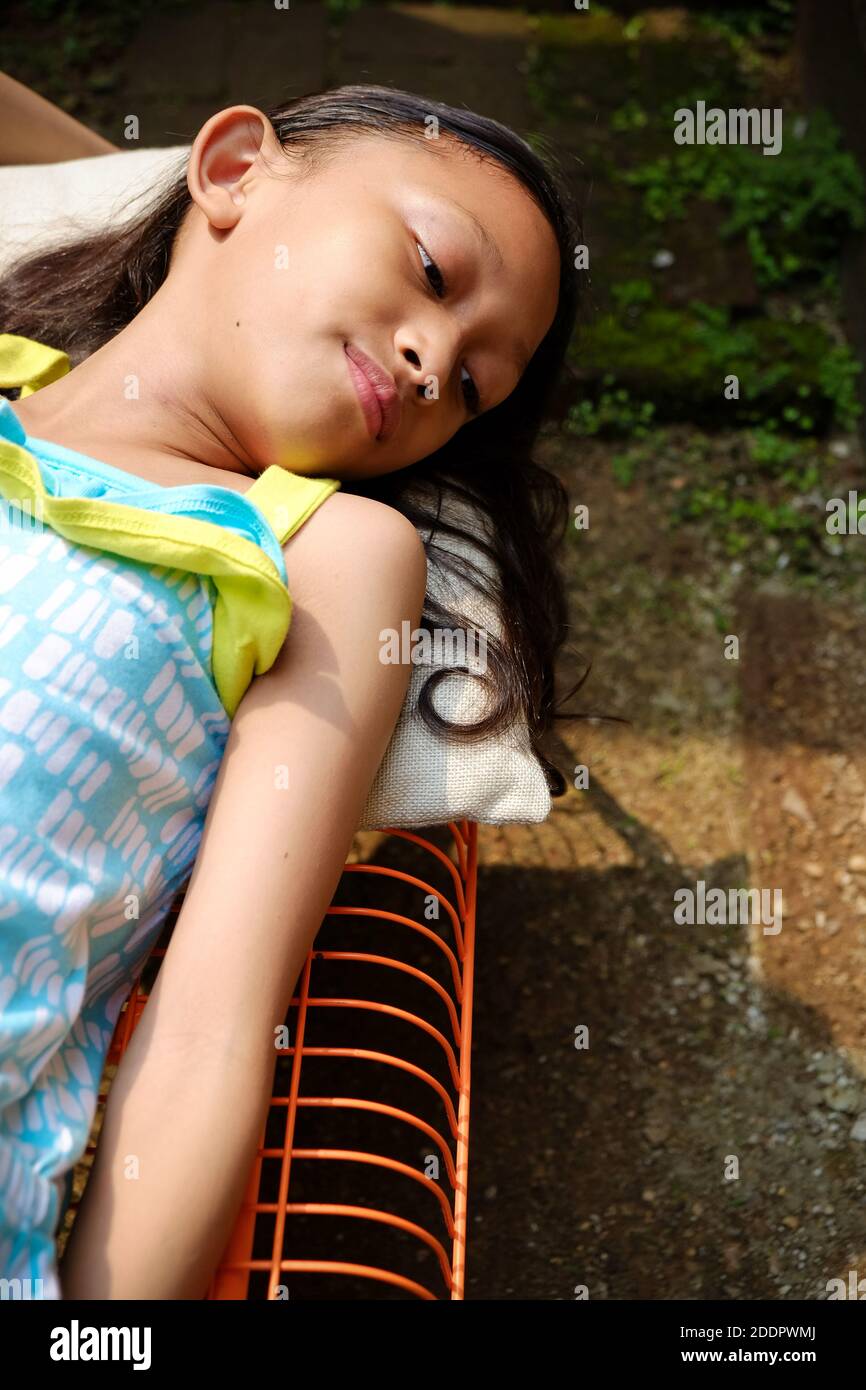Cute Asian ethnicity little girl lying down on a bench, sunbathing in the morning at home. hand rising up to cover her face from harsh sunlight Stock Photo