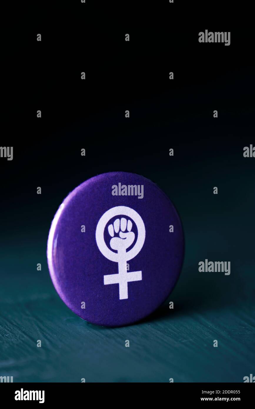 the women power symbol, a raised fist in a female gender symbol, in a violet pin button on a dark gray surface, against a black background with some b Stock Photo