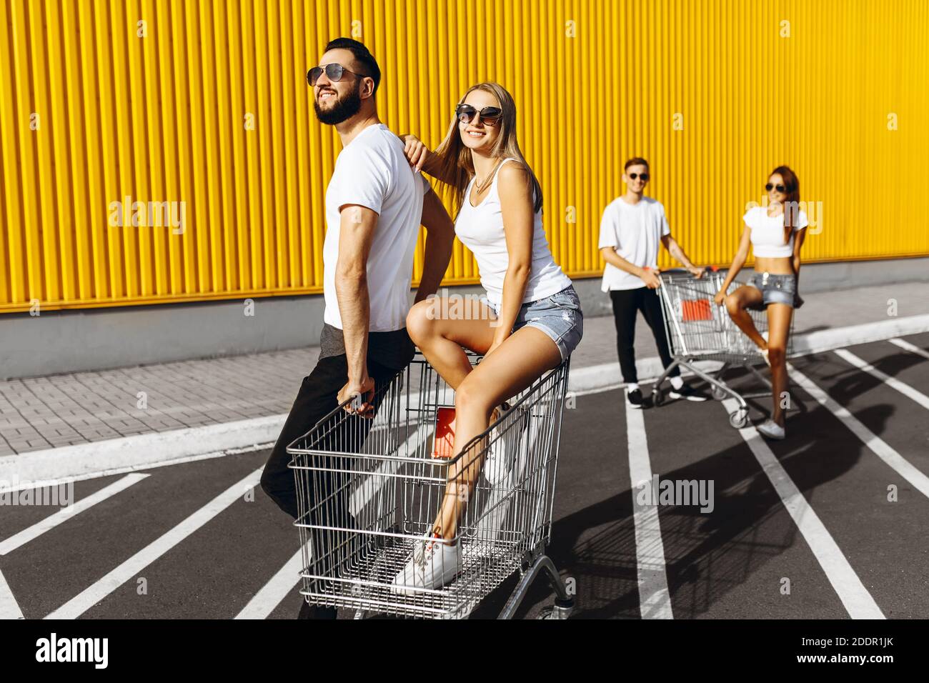 A group of young people, friends in white t-shirts, fun ride on carts near the store, supermarket, friends having fun, laughing in Sunny weather. Shop Stock Photo