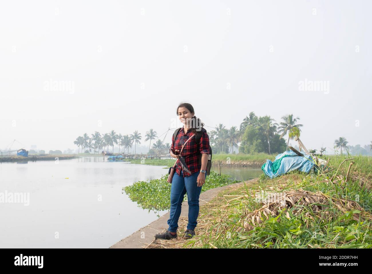 a Kerala women explorer with backpack on village road next to a river surrounded by green grass on white blur smoky background ,women power. Stock Photo