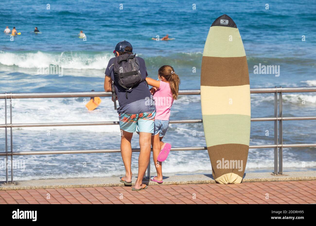 Adult and young girl standing next to surfboard looking out to sea. Stock Photo