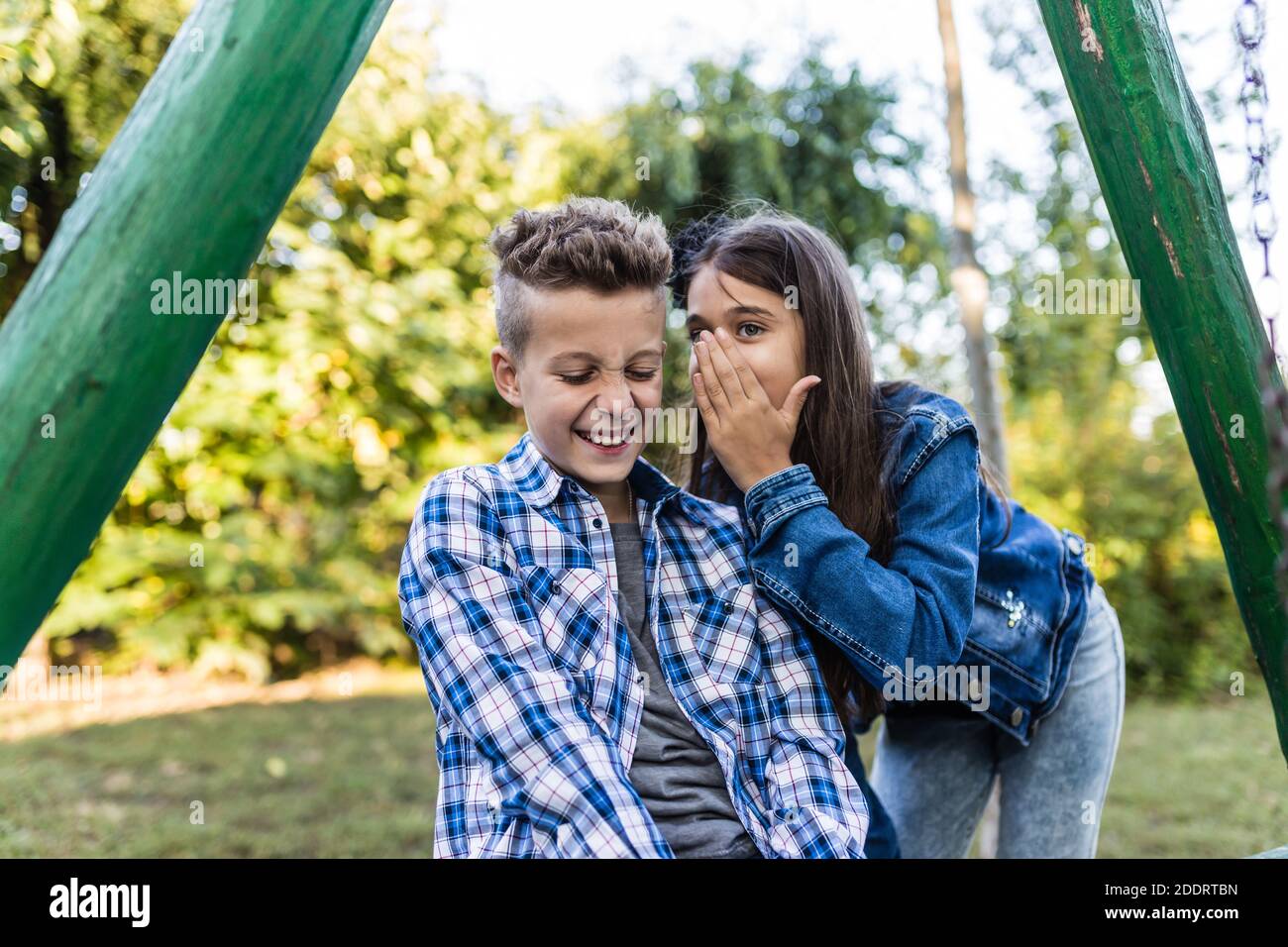 Close up two happy little friends  laughing and having fun in the park. Positive kids whispering a secret each other in the ears. Stock Photo