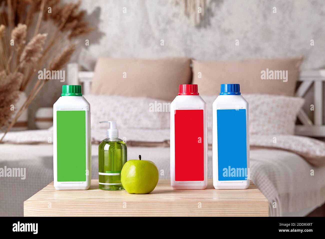 Three colorful plastic containers with detergents, pump bottle with soap and green apple on wooden bedside table in bedroom. Close up, copy space Stock Photo