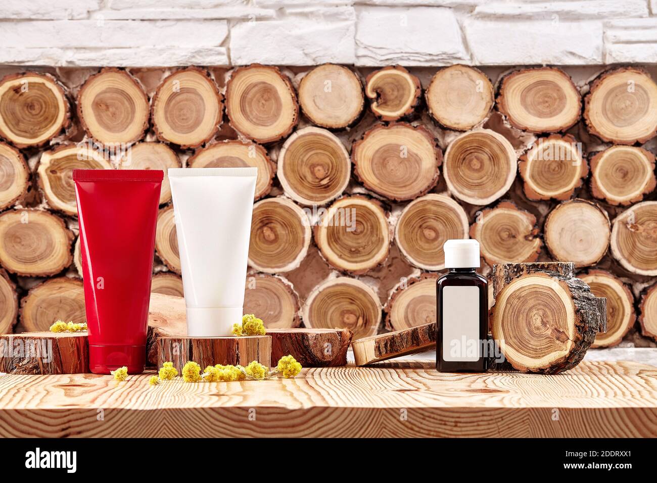 Two colorful cosmetic tubes and brown plastic bottle with branding area, small buds of blooming yellow flowers on wooden surface. Close up, copy space Stock Photo
