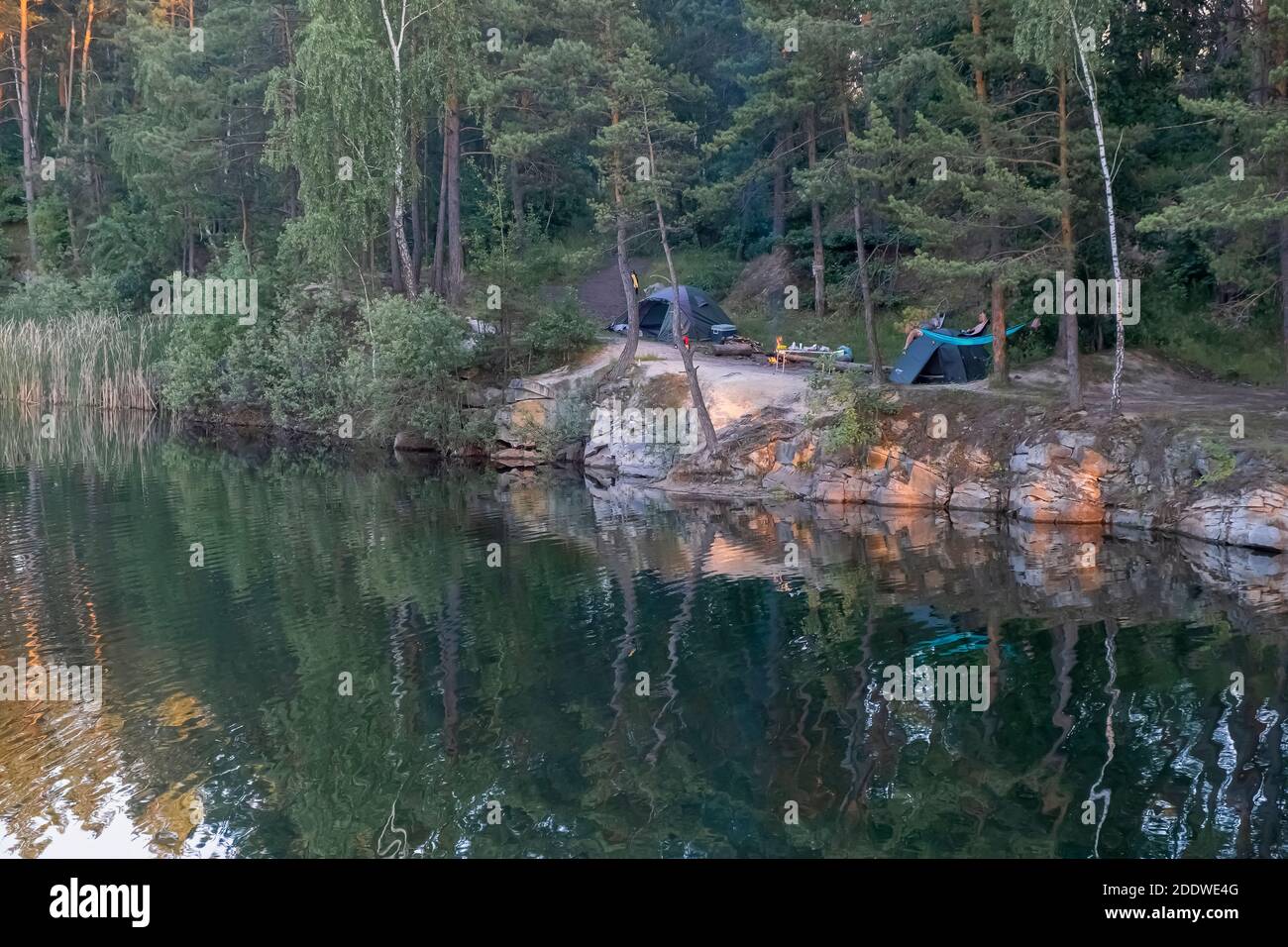 Camping on the lake. View of tourist tents located on the edge of a flooded granite quarry among pine trees Stock Photo