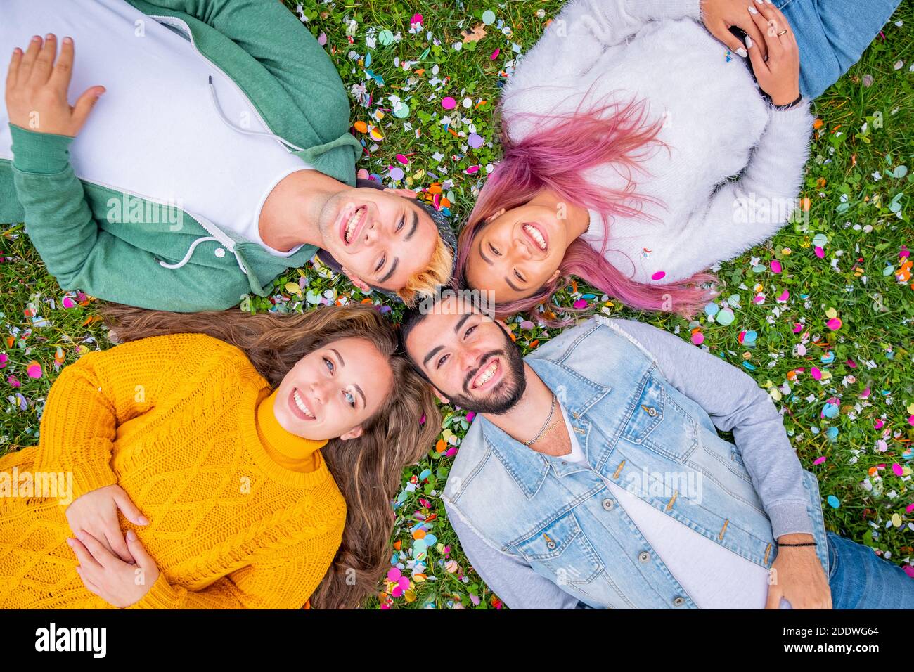 Group of smiling friends lying on the grass and colored confetti  outdoors   - Top view of beautiful stylish friends looking at camera in circle and s Stock Photo