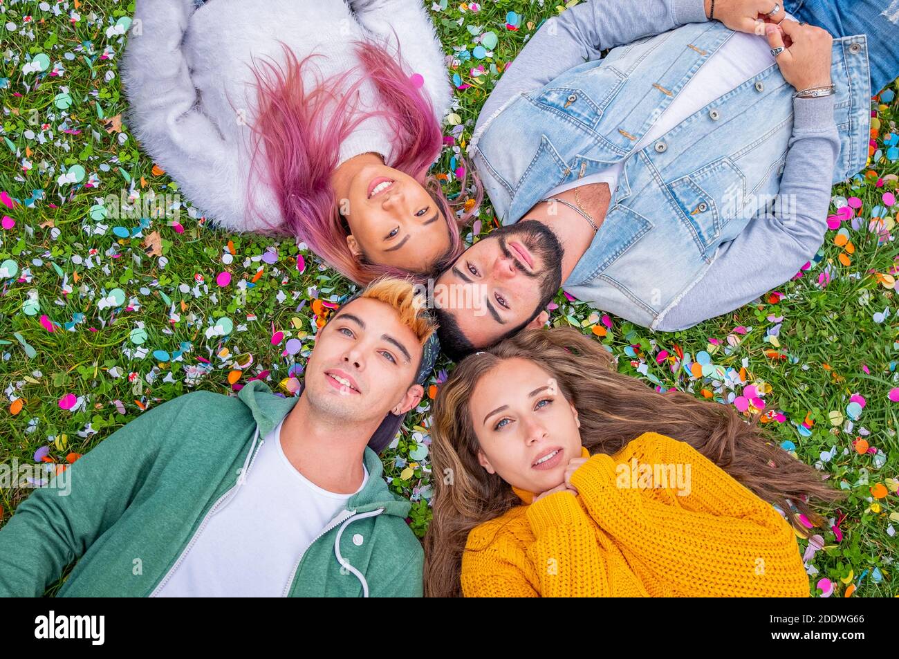 Group of smiling friends lying on the grass and colored confetti  outdoors   - Top view of beautiful stylish friends looking at camera in circle and s Stock Photo