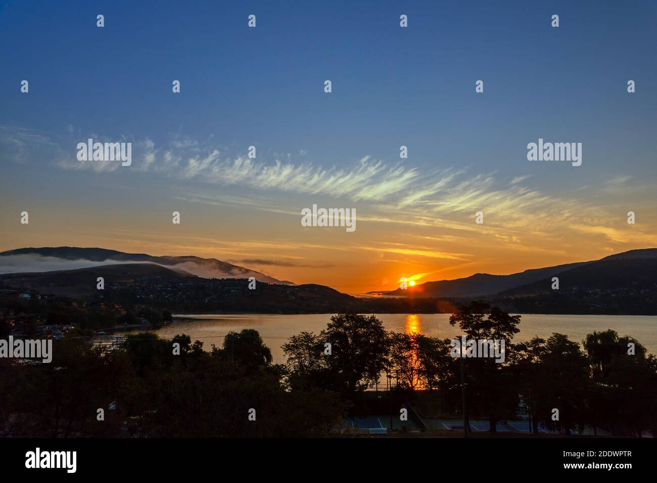 Sunrise over mountains and lake, near the coastal town. Haze of fog over the mountains, the reflection of the sun in the water, silhouettes of trees i Stock Photo