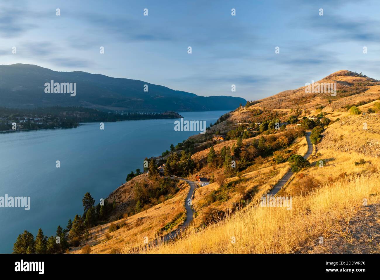 A hillside with yellow grass, paths, and private houses lit by the matins sun, the blue smooth surface of the river, silhouettes of mountains, and a b Stock Photo