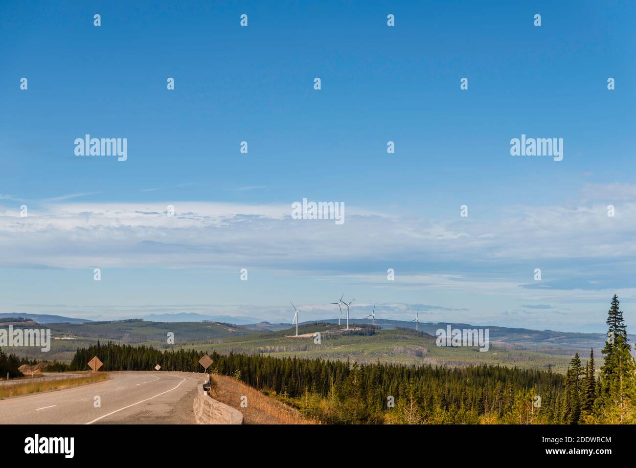 The view from the highway to the wind farms located on the hills covered with grass and trees. Cars are moving along the highway. Blue sky with clouds Stock Photo