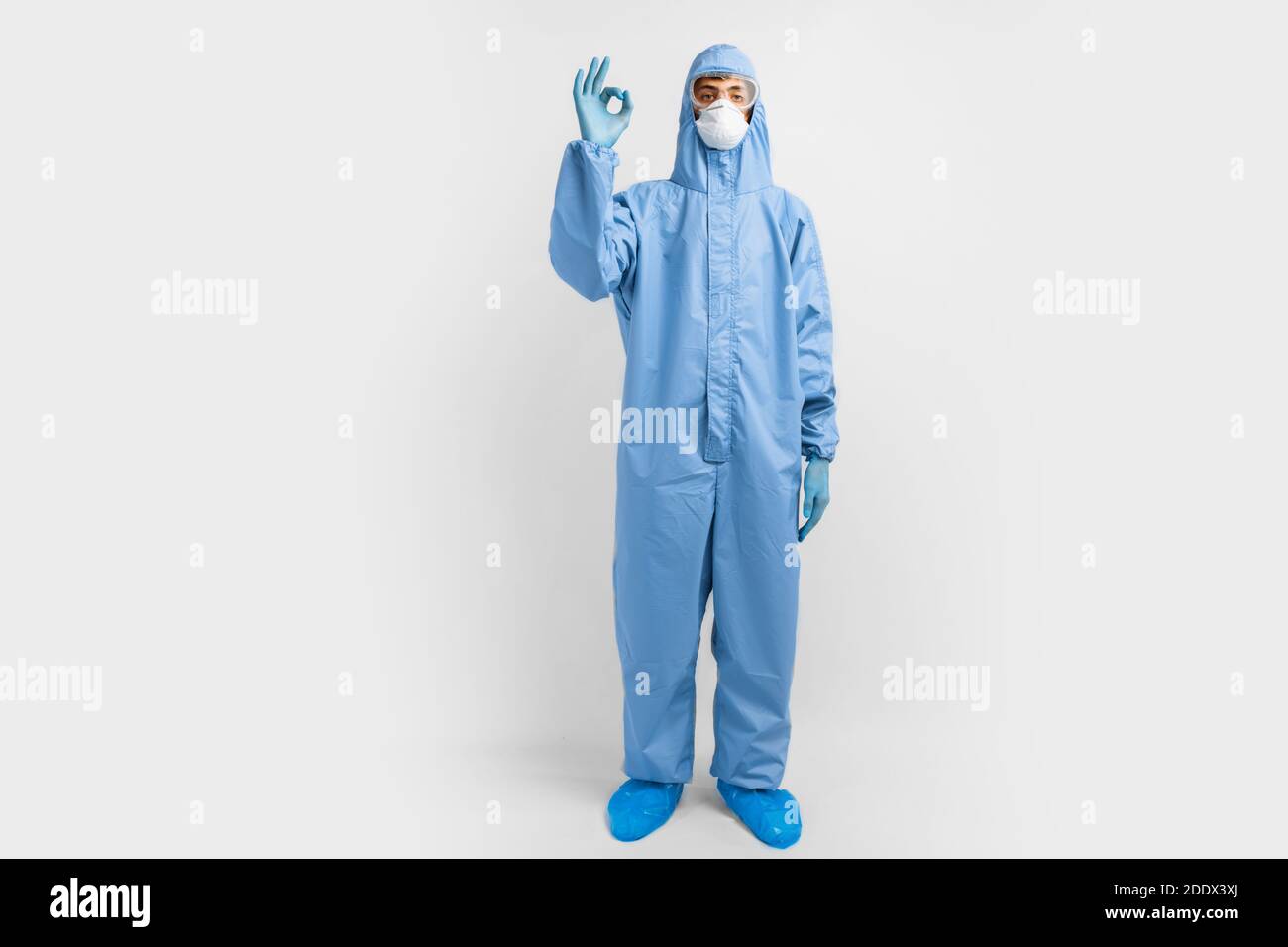 Positive man doctor, in protective blue suit, glasses, mask and gloves, shows okay gesture, on white background, healthcare, coronavrus Stock Photo