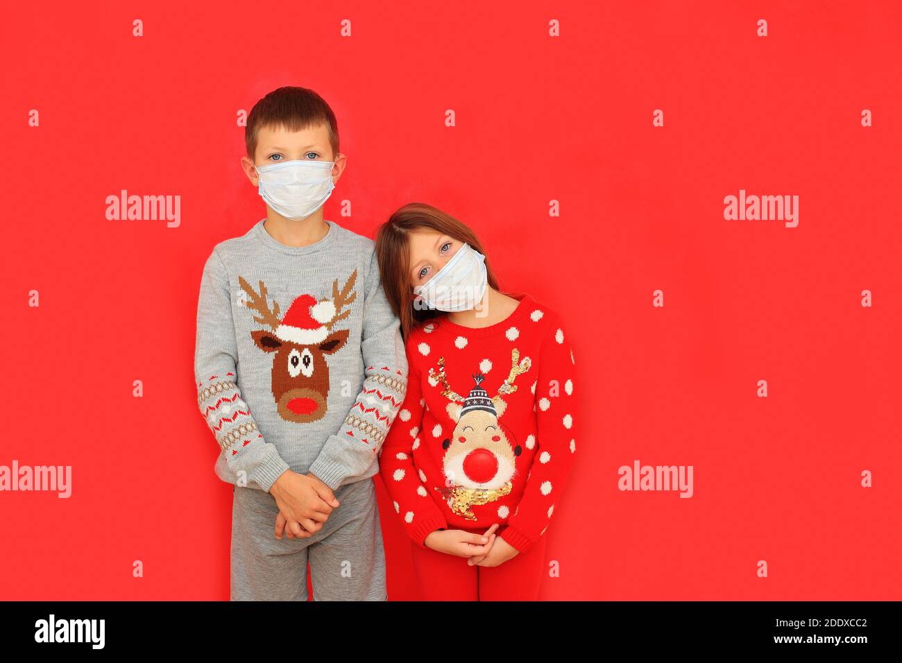 A boy and a girl wearing respiratory masks keep their distance on New Years during the quarantine period. Children in New Year's knitted sweaters on a red background Stock Photo