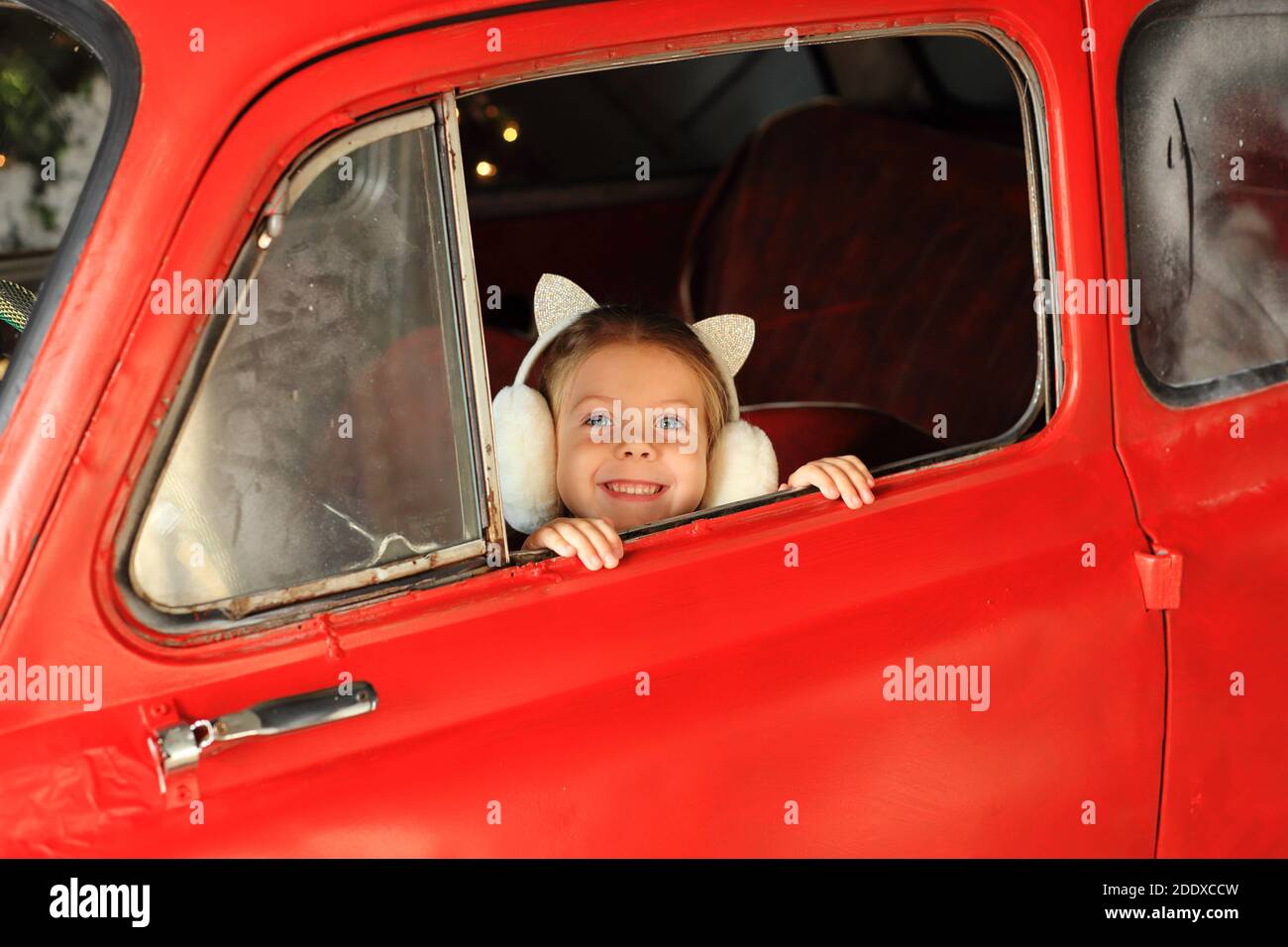 A little girl is sitting in a red car with New Year's gifts. The child looks out of the car Stock Photo