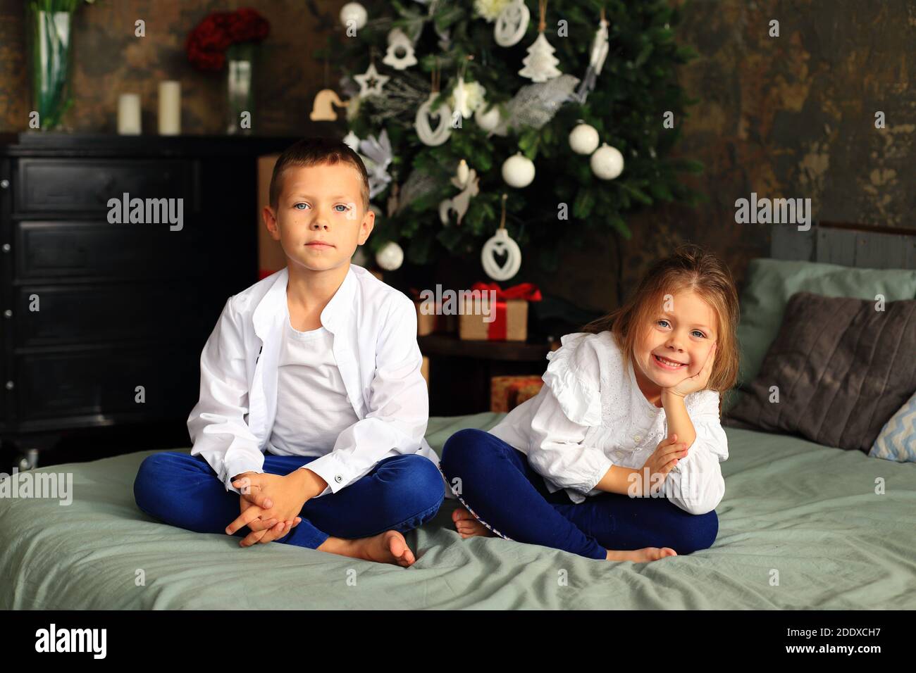 Children are sitting on the bed in front of the Christmas tree. Boy and girl waiting for christmas Stock Photo