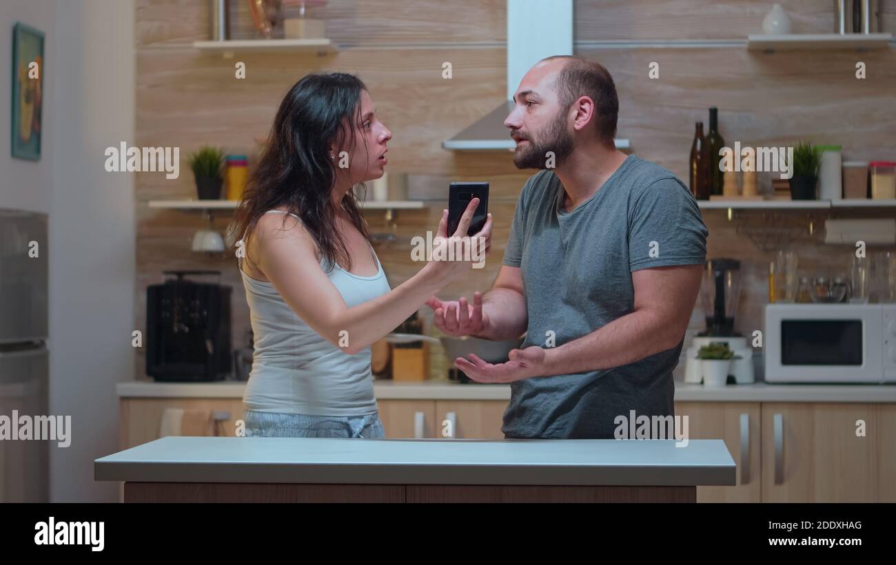 Wife screaming at unfaithful husband in the kitchen. Jealous woman cheated angry frustrated offended irritated accusing her man of infidelity showing him messages on smartphone yelling desperate. Stock Photo