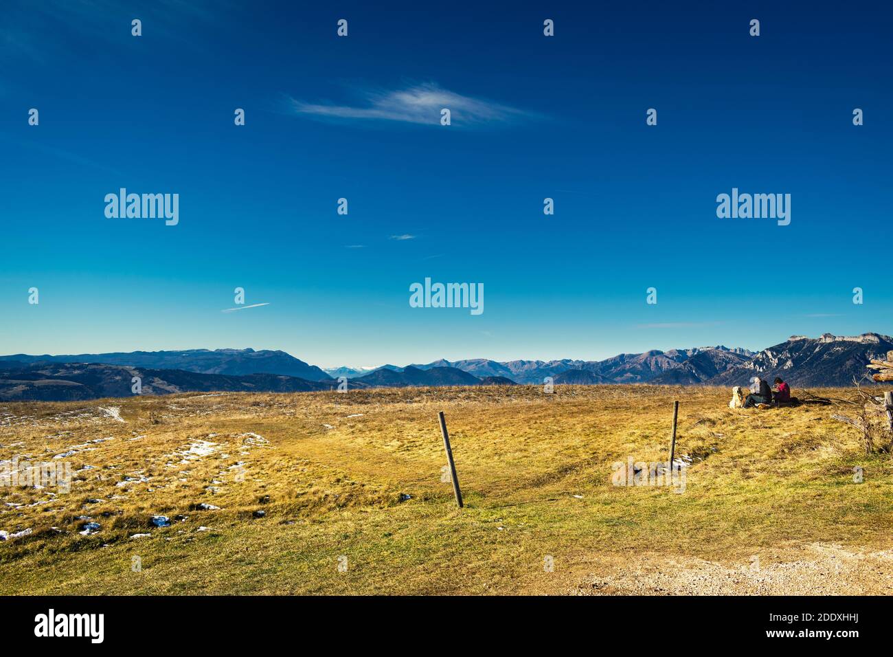Winter panorama from the top of Monte Avena. Clear skies and mountain ranges in the distance. Couple of tourists with dog rests on the lawn. Stock Photo