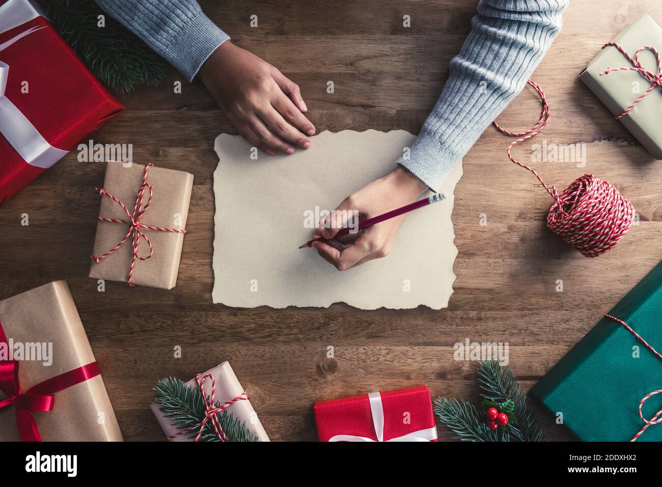 Woman in a gray sweater writing on craft paper with a red pencil in the center of christmas gifts on a wooden table Stock Photo