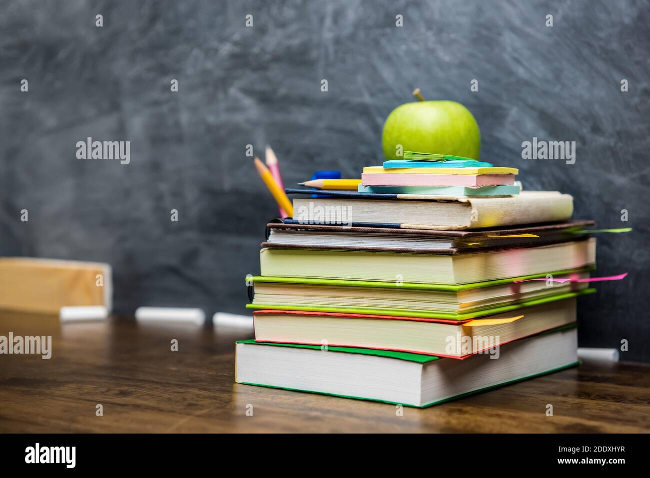 Stack of books, stationery and education supplies on wooden table in classroom with blackboard in background Stock Photo