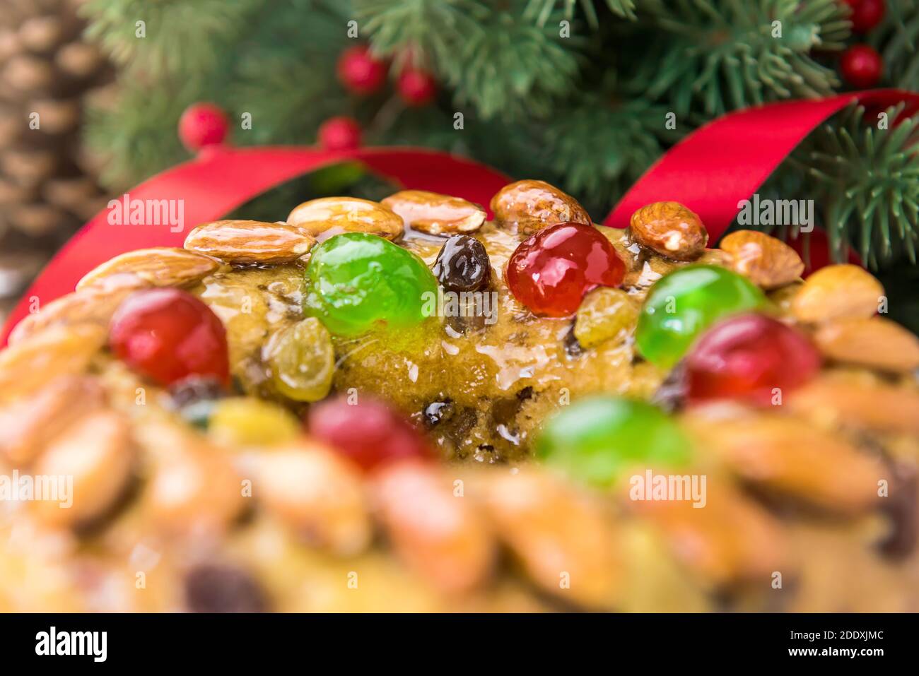 Close up of glazed round hollow colorful Christmas fruitcake with green pine needles and red ribbon decorations in background Stock Photo