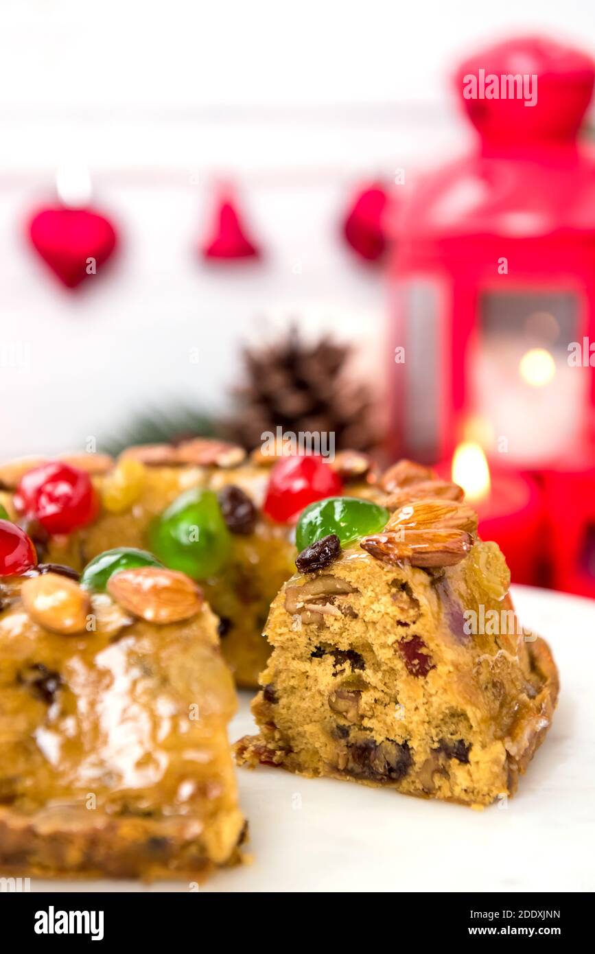 Glazed colorful Christmas fruitcake topped with almonds and glace cherries on white platter with candles and decoration items in background Stock Photo