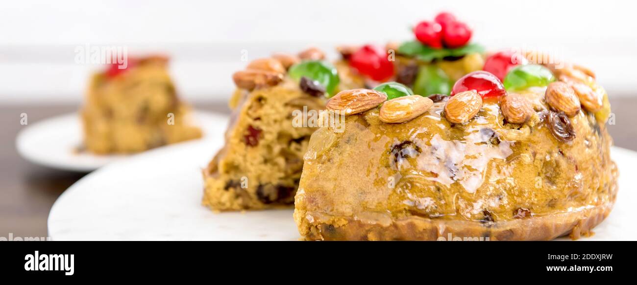 Close up of glazed colorful Christmas fruitcake topped with almonds and glace cherries on white platter, panoramic banner Stock Photo
