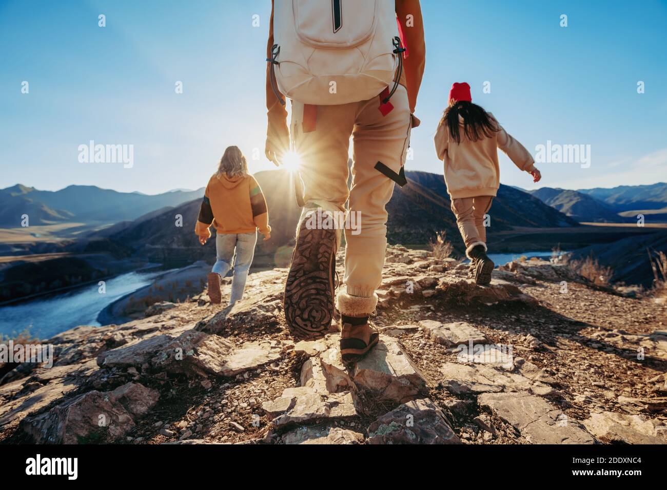 Three young tourists hikers walks against sunset mountains and river Stock Photo