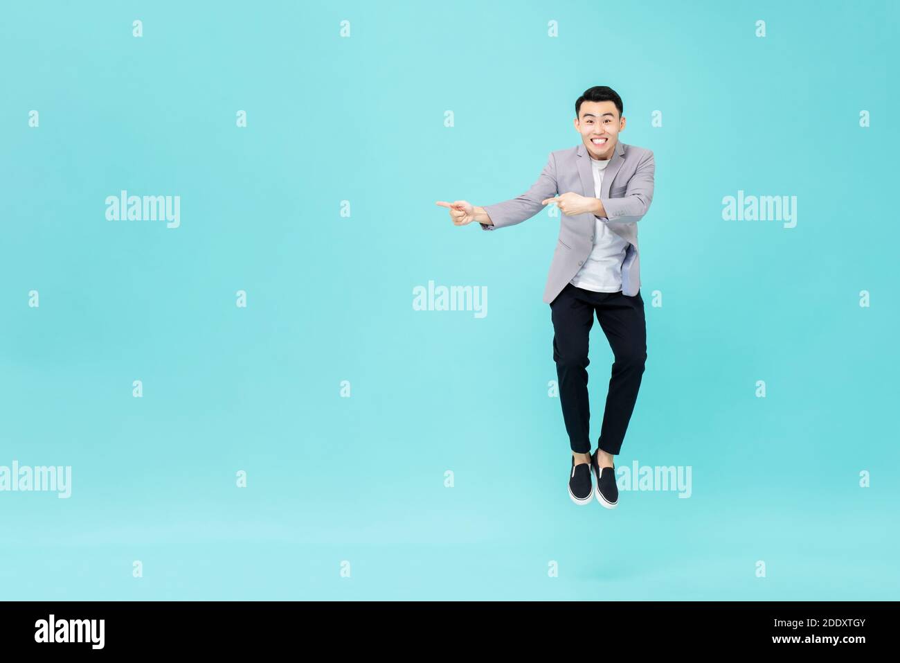 Young smiling Asian man with funny face jumping and pointing hands to copy space aside on studio light blue background Stock Photo