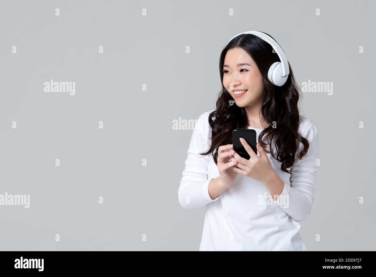 Asian woman in white t-shirt wearing wireless headphones listening to music from smartphone isolated on light gray background Stock Photo
