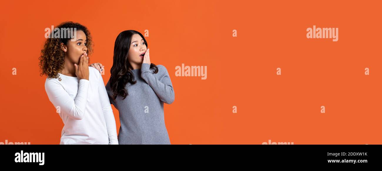 Asian and African American woman friends in shocked gesture with hands cover mouths isolated on orange banner background with copy space Stock Photo