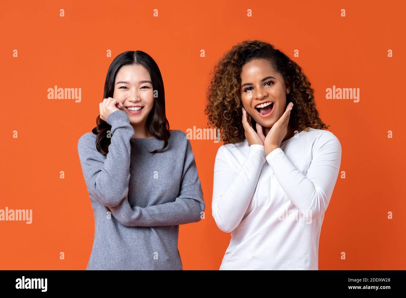 Smiling Asian and African American woman friends in surprised gesture isolated on orange background. Stock Photo