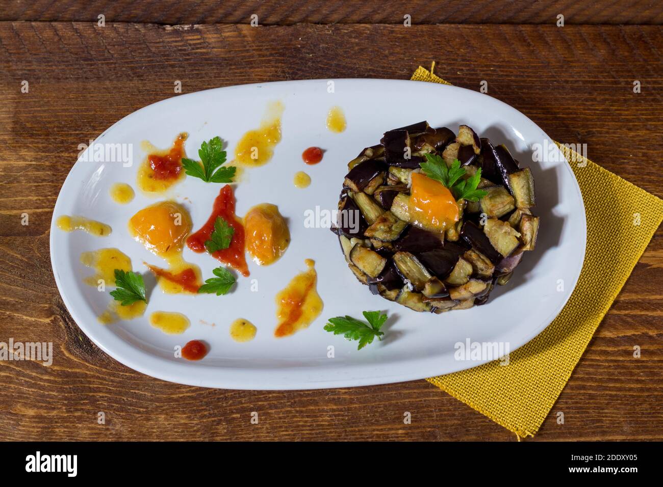 Plate of diced aubergines on a cream of yellow and red cherry tomatoes in a white plate. Wood background. Selective focus. Stock Photo